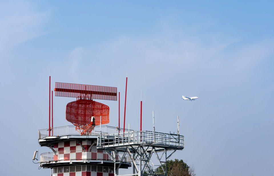 Torre de radar ao lado de um avião contra o céu azul, representando o monitoramento do espaço aéreo.