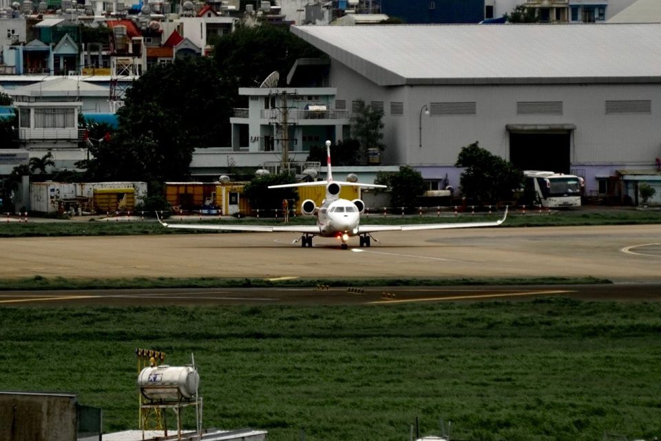Jato executivo alinhado à pista de um aeroporto, com edifícios urbanos ao fundo.