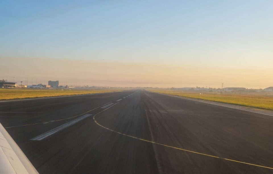 Pista de aeroporto vazia ao amanhecer, com edifícios aeroportuários ao fundo.