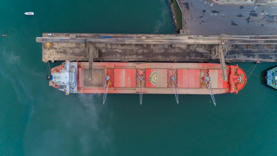 Navio de carga atracado em um porto brasileiro, visto de cima, com áreas de operação e acesso marítimo ao redor.